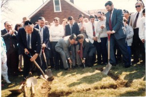 A crowd of men in suits gathered on a lawn, several of them turning earth with shovels.